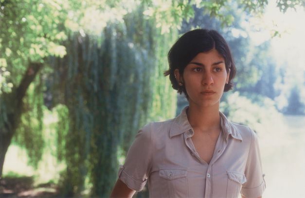 Film still from DER SCHÖNE TAG: A woman with chin-length hair looks to the right. There are trees in the background. The sun is shining.