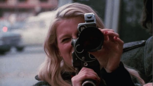 Filmstill aus PLACE DE LA REPUBLIQUE: Eine Frau hält dem Betrachter eine Kamera ins Gesicht. Hinter ihr eine Straße und Autos.