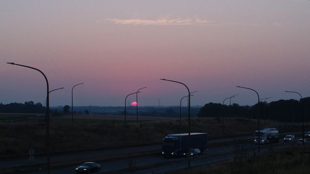Filmstill aus dem Film „hold on to her“ von Robin Vanbesien. Die rosafarbene Sonne geht im Hintergrund einer scheinbaren Landstraße unter, und ausgeschaltete Straßenlaternen füllen den Rahmen.