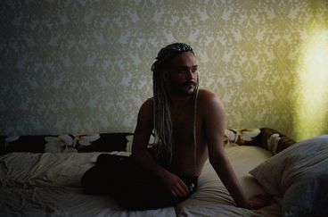 A shirtless man with braids sits on a futon, looking to the right of the frame. Behind him we see patterned wallpaper.
