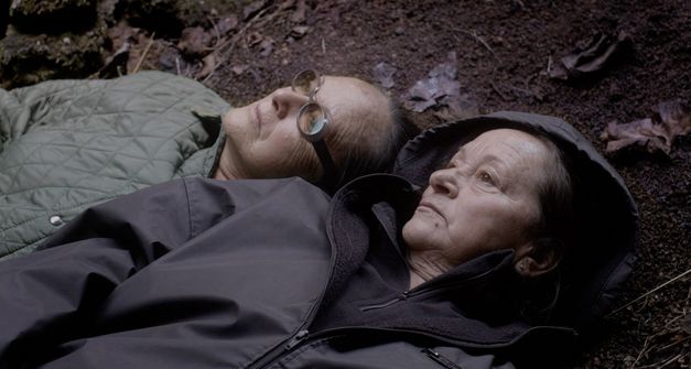 Film still from "La hojarasca" by Macu Machín. It shows two elderly people lying on the forest floor and looking up. 
