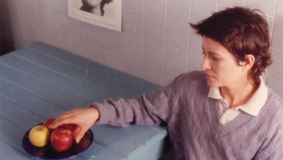 Filmstill aus UM ADEUS PORTUGUÊS: Eine junge Frau sitzt an einem Tisch. Sie greift nach den Äpfeln, die in einem Teller auf dem Tisch stehen.