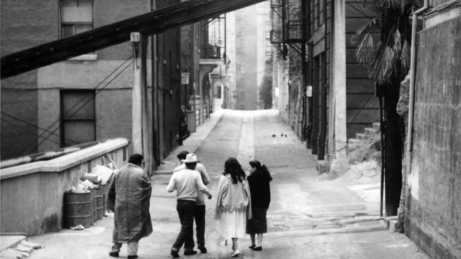 Film still from THE EXILES: A group of people walk along an otherwise empty street.