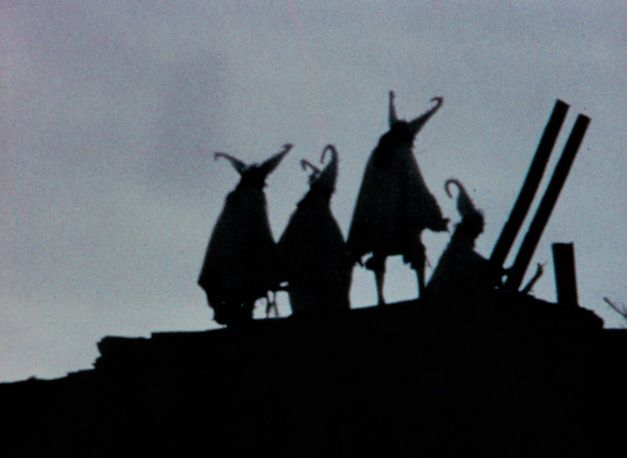 Film still from "Techqua Ikachi, Land – mein Leben" by Anka Schmid, James Danaqyumptewa and Agnes Barmettler. It shows a gray sky and dark figures standing on a hill. The people are all wearing pointed hats. 
