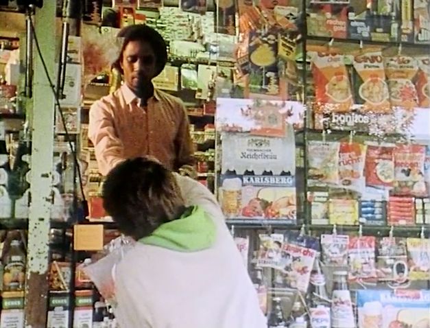 Still from the film „Man sa yay“ by Safi Faye. It shows a shop for food and tobacco products behind glass. A man in the shop stands at an open window and hands something to a person.