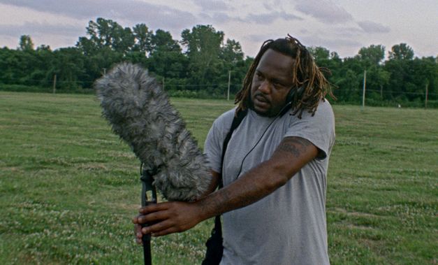 Film still from Theresa Delsoin, Lisa Malloy, JP Sniadecki, Ray Whitaker’s film “On the Battlefield”. A sound recordist stands in a field holding a boom pole and wearing headphones, and listens intently to what they are recording. In the background we see a cloudy sky and trees. 