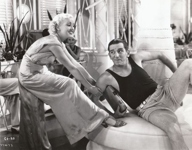 Film still from THE GAY DIVORCEE: Edward Everett Horton is sitting on a sofa in shorts and a tank top, a young woman playfully tugging at him.