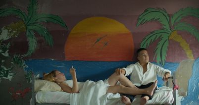 Still aus dem Film REAS von Lola Arias (2023): Eine Frau und eine Mann auf einem Bett. Dahinter ein Wandbild eines Sonnenuntergangs mit Palmen. 