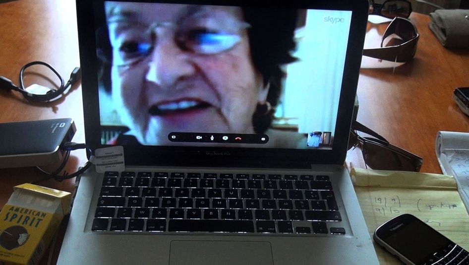 Filmstill aus NO HOME MOVIE: Auf einem Tisch steht ein Laptop, auf dem man eine ältere Frau beim Skype-Gespräch sieht.