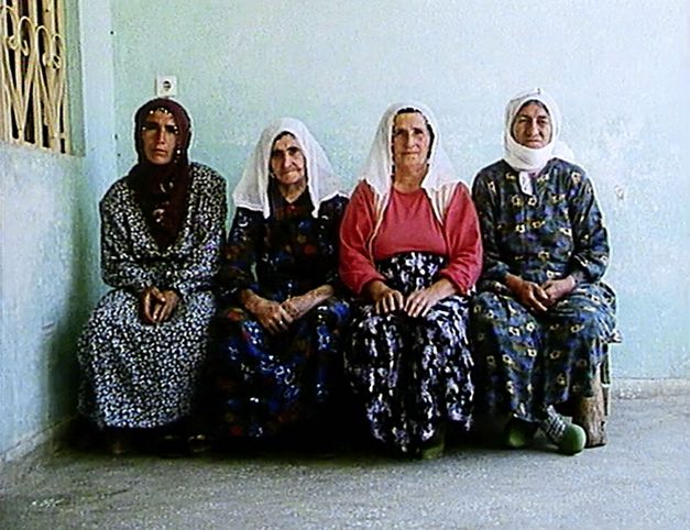 Still from the film „Mein Vater, der Gastarbeiter“ by Yüksel Yavuz. Four women in headscarves sit next to each other on a bench against the wall of a house.