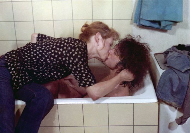 Film still from RUMMEL: A man lies in the bathtub. A woman sits on the edge of the bathtub and kisses the man. Towels and clothes can be seen on the edge.