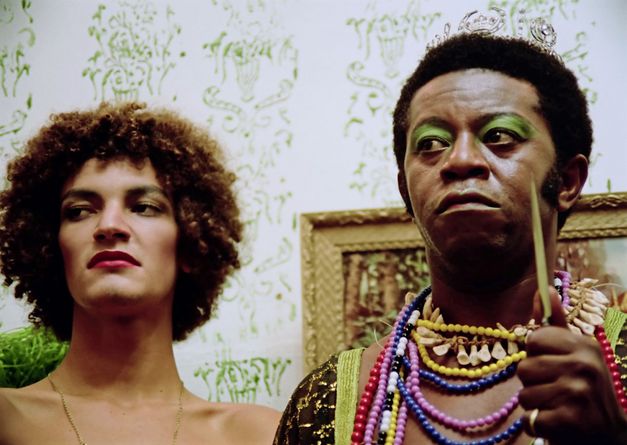 A close-up of two men wearing make-up. The man on the left is not wearing a top, but only a long narrow necklace. The man on the right wears many colourful necklaces and holds a knife in his hand. 