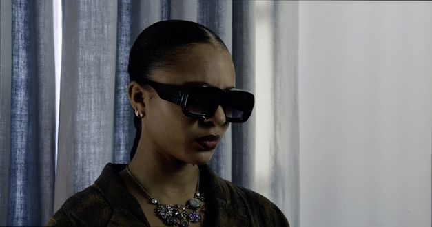 Film still from "The Wrong Movie" by Keren Cytter. It shows a close-up of a woman with braided hair, a sparkling necklace and large black sunglasses. Tears are running down her cheek.