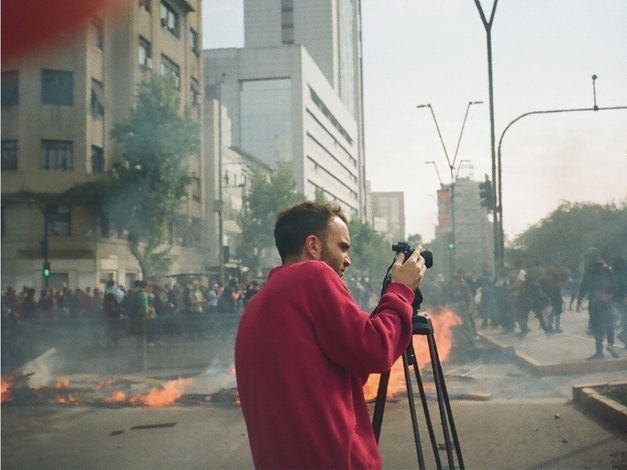 35mm colour photo of a man in a red sweater on a city street looking into a camera on a tripod. Behind objects are on fire, and further in the background is a crowd of people 