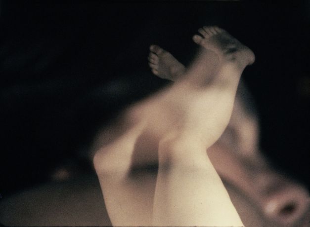 Film still from "Iris" by Maria Lassnig. It shows two naked legs stretched up in the air. The image is overlaid with a close-up of a person.