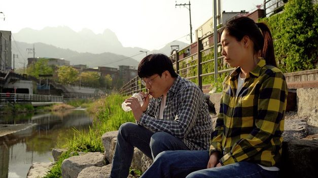 Filmstill aus „Hot in Day, Cold at Night“ von Park Song-yeol. An einem sonnigen Tag sitzen ein Mann und eine Frau an einem Bach. Der Mann trinkt etwas mit einem Strohhalm aus einem Plastikgefäß.