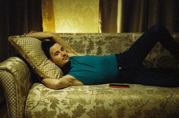 A man in a blue, sleeveless shirt and black pants lounges on a couch, with his hands stretched over his head. His phone is beside him. 