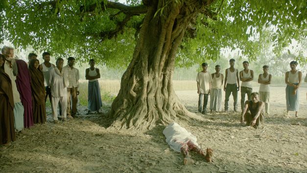 Filmstill from "In the Belly of a Tiger" by Siddartha Jatla. It shows a tree, with people standing to the left and right of it, looking towards the camera. Under the tree lies a person wrapped in a white cloth. 