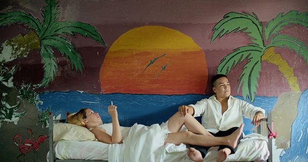 Film still from "Reas" by Lola Arias. It shows a smoking woman lying on a bed and a man sitting at the foot of the bed. The wall is painted with a sunset.