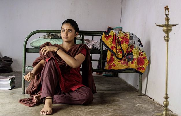 Filmstill aus "Kottukkaali" von Vinothraj PS. Zu sehen ist eine Frau mit geflochtenem Haar und einem Punkt zwischen den Augenbrauen, die mit dem Rücken zu einem Bett auf dem Boden sitzt. 