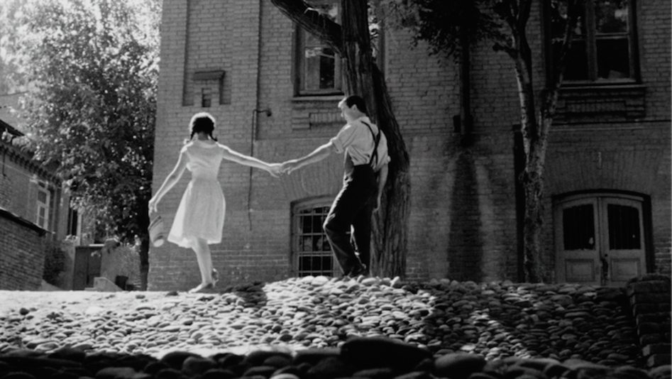 Filmstill aus APRILI: Auf einer gepflasterten Straße halten sich zwei junge Menschen an den Händen.