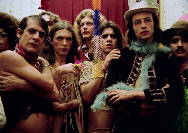 A group of men can be seen, all looking towards the camera. They are wearing make-up and colourful, eye-catching accessories. In the middle of the picture, a festoon hangs down from the ceiling. 