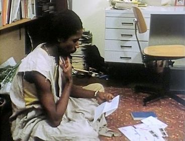 Still from the film „Man sa yay“ by Safi Faye. A Man sits on the floor in an office reading a letter. He is surrounded by office furniture and documents.