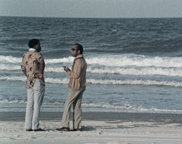 Still from the film "I Heard It through the Grapevine" by Dick Fontaine. Two men, one of them wearing black sunglasses, are standing on a sand beach and looking at each other, seemingly in conversation. Behind them, waves are washing up from the sea.