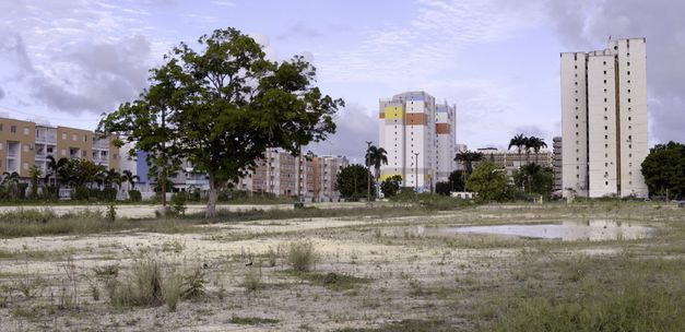 Film still from "L’ homme-vertige" by Malaury Eloi Paisley. It shows a green space with a few trees, sand and a large puddle next to a town. 