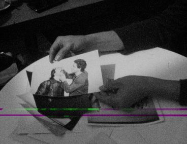 Still from the film "This Makes Me Want to Predict the Past" by Cana Bilir-Meier. A close-up of two hands holding a photo. The photo shows two men, one of them holds the head of the other so that the latter looks up. The former looks intently at his face. 