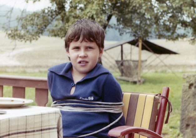 Film still from VERSTECKTE FALLEN: A tied-up boy sits at a garden table.