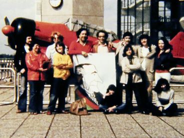 Filmstill from „Aufenthaltserlaubnis" by Antonio Skármeta. Old photo of a group of people posing with a caricature of the Shah of Iran in front of a small red plane.
