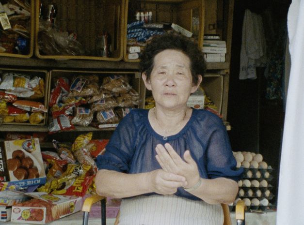 Film still from "Doesarananeun moksori / Yomigaeru Koe" by Park Soo-nam and Park Maeui. It shows an elderly lady in a blouse sitting on a chair and looking towards the camera. Behind her are shelves full of groceries. 