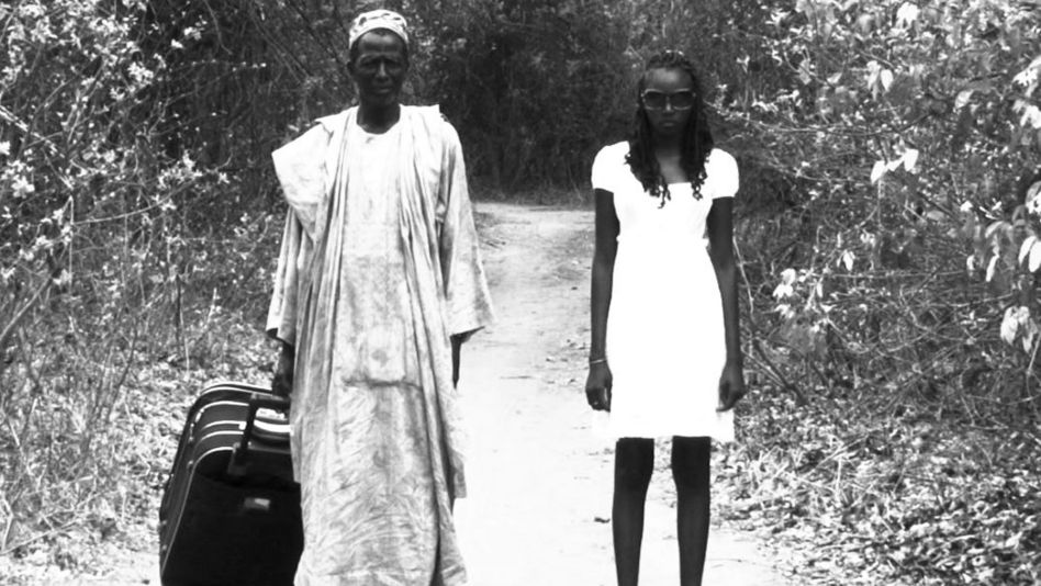 Film still from A BATALHA DE TABATÔ: A man and a younger woman stand next to each other on a country lane and look towards the camera. He is pulling a suitcase behind him.