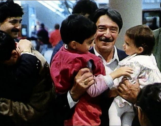Still from the film „Mein Vater, der Gastarbeiter“ by Yüksel Yavuz. A man with a moustache holds two children in his arms and smiles. On the left side of the picture, two people are hugging each other. Several people are standing in the background. 