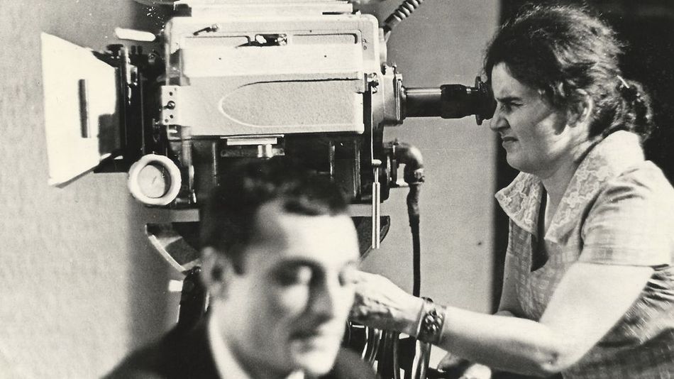 Film still from "Deda-Shvili an rame ar aris arasodes bolomde bneli" by Lana Gogoberidze. It shows a black and white image of a woman using a video camera. 