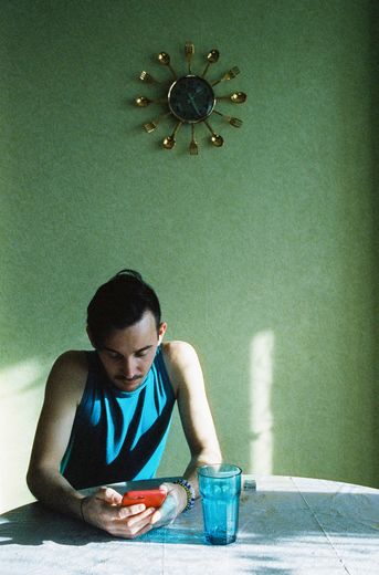 A man in a blue, sleeveless shirt sits at a sparse table and looks down at a phone in a red case. Behind him is a clock on a greenish wall.