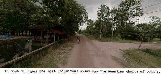 Filmstill aus dem Film „Trip After“ von Ukrit Sa-nguanhai. Eine Straßenansicht in einer bescheidenen ländlichen Gegend. In der Mitte des Bildes ein*e Radfahrer*in. In der Bildunterschrift heißt es: „In most villages the most ubiquitos sound was the unending chorus of coughs.“