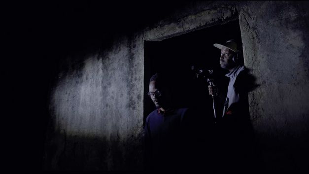 Film still from "The Nights Still Smell of Gunpowder" by Inadelso Cossa. It shows two men in a dark door frame.