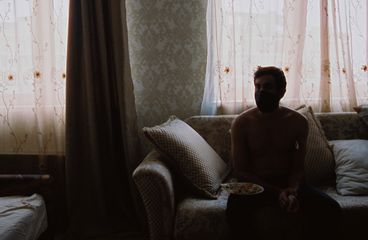 Ein oberkörperfreier Mann mit einer schwarzen Maske sitzt auf einer Couch und hält einen Teller mit Essen. Er ist teilweise in Silhouette, das Licht durch blumige, gemusterte Vorhänge gefiltert. 