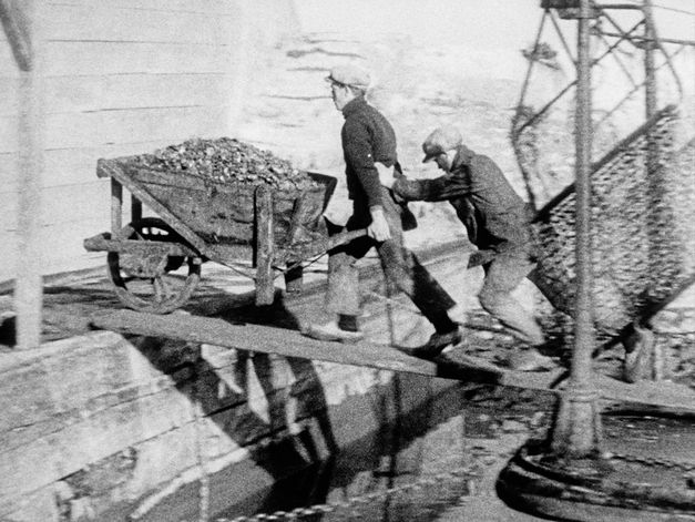 Filmstill aus „Dearest Fiona" von Fiona Tan. Altes Bild in Schwarzweiß von zwei Arbeitern, die eine Schubkarre auf einem Balken über das Wasser schieben.