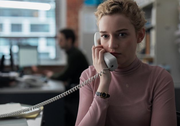 Film still from THE ASSISTANT: A young woman with blonde hair sits in an open-plan office and holds a telephone to her ear.