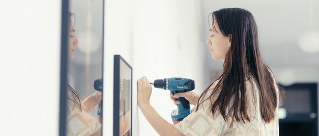 Film still from "Reproduktion" by Katharina Pethke. It shows a woman working with a drill on a white wall.