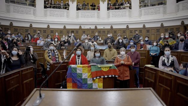 Film stil from "Oasis" by Tamara Uribe and Felipe Morgado. It shows a full courtroom. The people in the courtroom are wearing medical masks and looking towards the camera. 