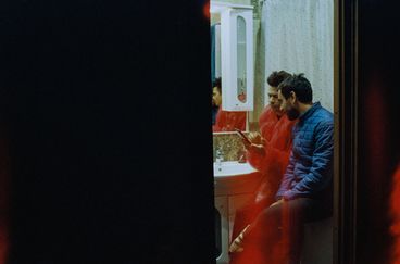 A mostly black composition, with a vertical sliver that reveals two men in a bathroom leaning backwards against a bathtub as one shows the other something on a mobile phone. On wears a red jacket, the other a blue jacket. 