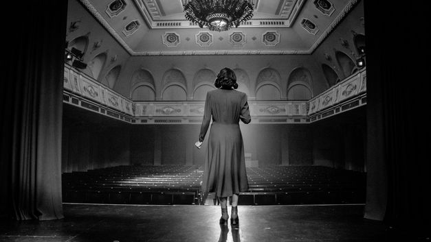 Film still from "Marijas klusums" by Dāvis Sīmanis. It shows a woman on a theatre stage, standing with her back to the camera and looking into the empty theatre hall. 