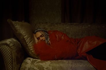 A man with braids, wearing a red fur and leather jacket, appears to sleep on a couch in a darkened room.