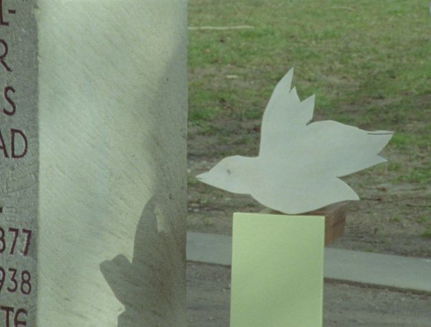 Filmstill aus dem Film „Zwischenwelt“ von Cana Bilir-Meier. Auf der linken Seite sehen wir ein Teilstück eines Denkmals aus Stein. Auf der rechten Seite ein Vogel aus Holz. 