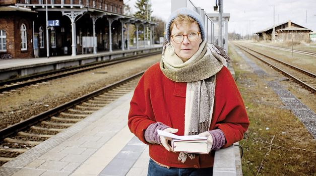 Still from the film "Gehen und Bleiben" by Volker Koepp. A person wearing a blue hat, a beige scarf, a red jacket and red glasses is standing on a railway platform. They are holding a book with both hands.