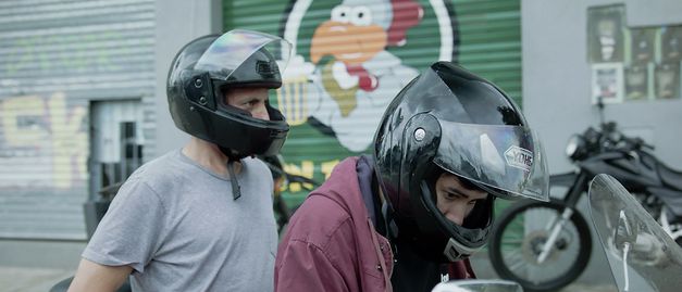 Still from the film "Camuflaje (Camouflage)" by Jonathan Perel. Two men with helmets on share a motorbike. 